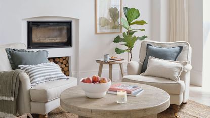 A neutral living room with two large white arm chairs and an unlit fireplace, firewood stacked beneath
