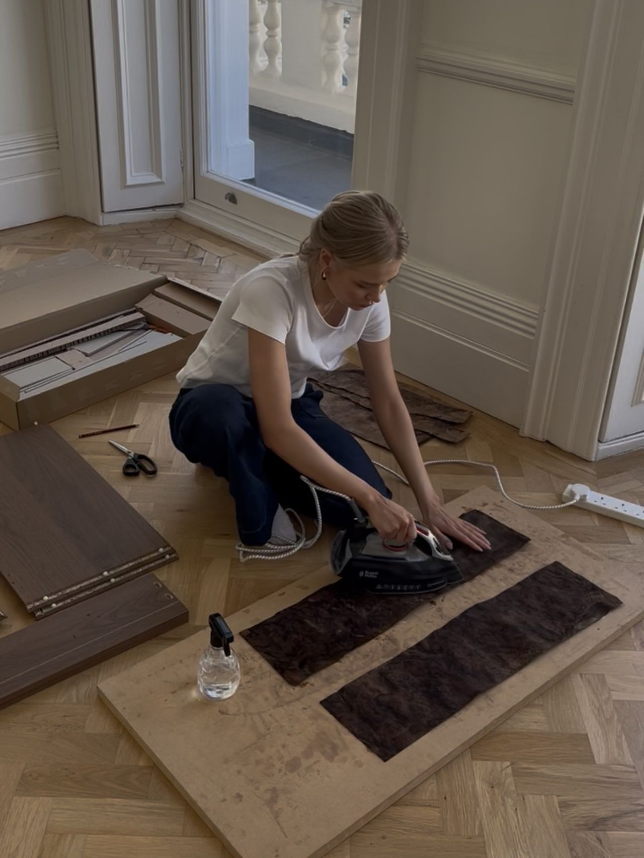 Image of a woman in a white t-shirt and jeans, ironing a dark piece of burl wood vinyl onto a wooden board that is lying on a wooden floor.