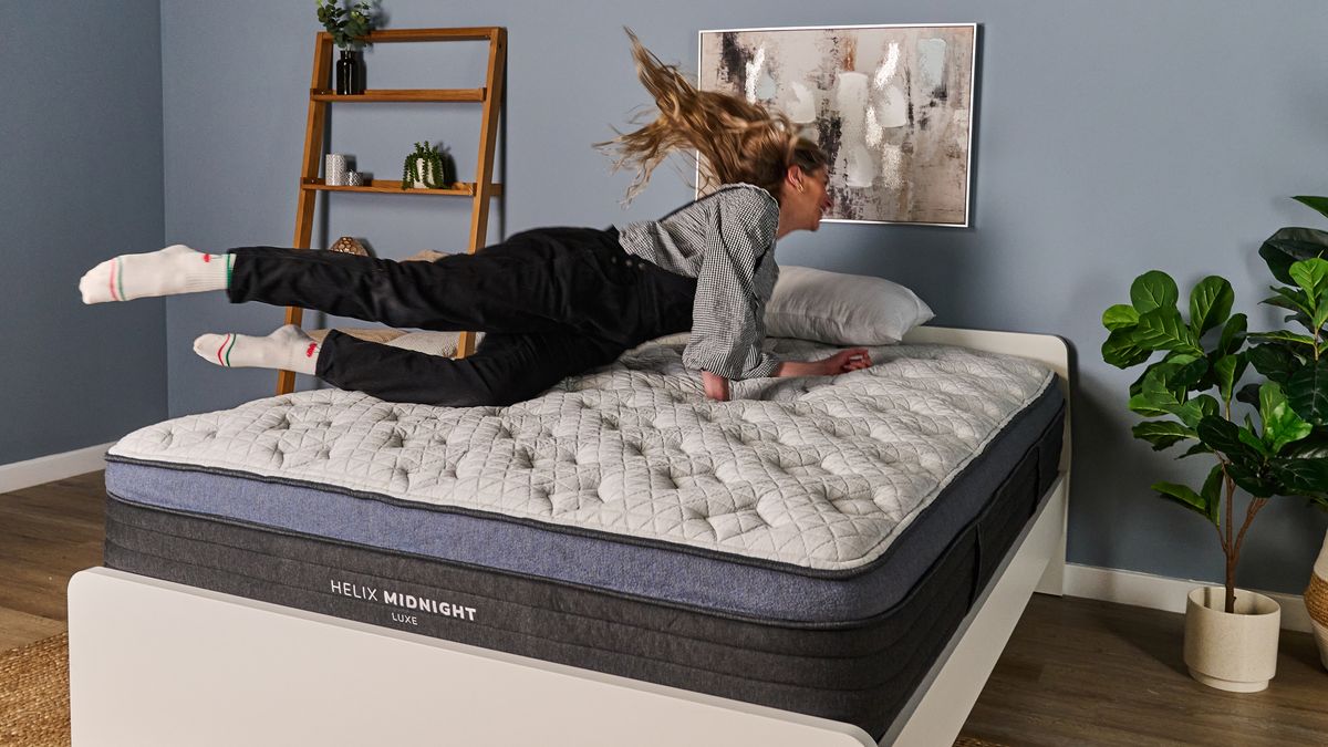 The image shows a woman diving onto a white pillow top mattress with blue trim. The mattress is on a white bed frame in a blue bedroom.