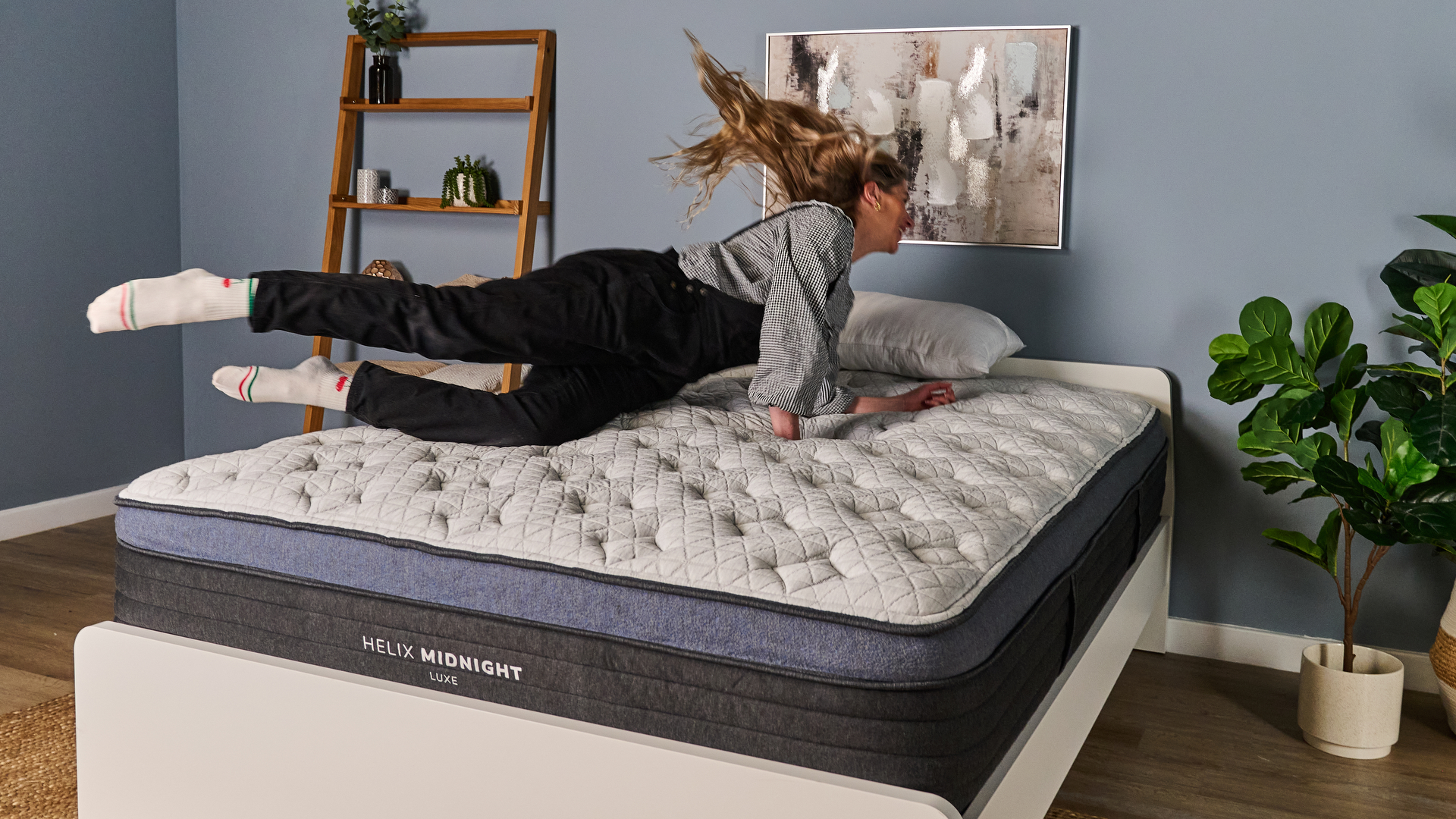 The image shows a woman diving onto a white pillow top mattress with blue trim. The mattress is on a white bed frame in a blue bedroom.