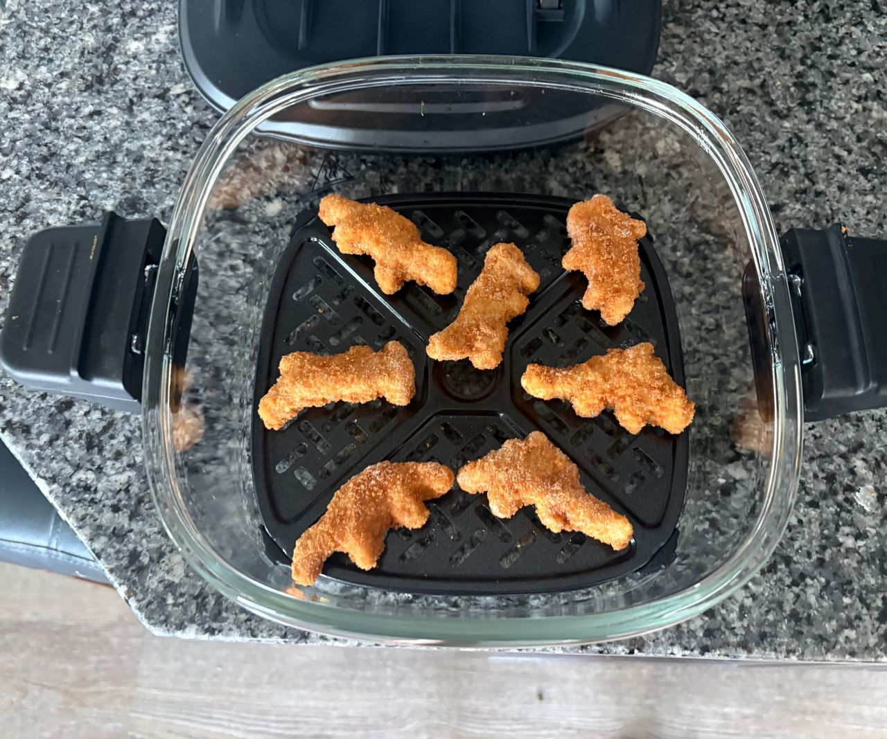 The Cuisinart glass air fryer on a granite countertop, viewed from above with dino nuggets inside.