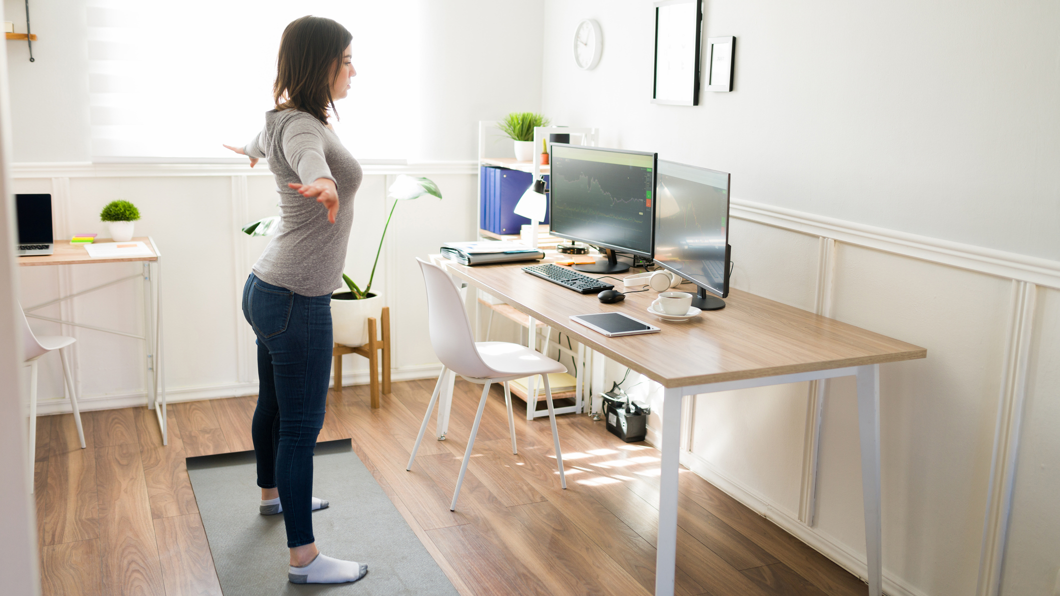 Woman stands on yoga mat with arms outstretched, looking at computer screens on a desk