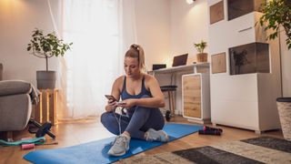 A woman looking at her workout plan on her phone sat on the living room floor