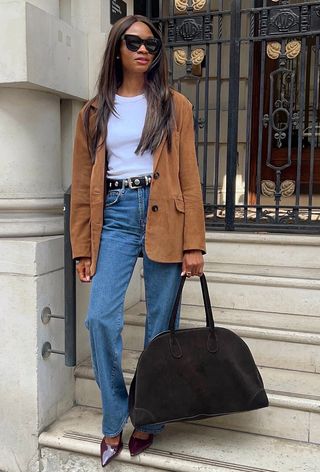 best elevated basics shown in a photo of a woman wearing a tan suede jacket over a white t-shirt with a studded belt, jeans, pointed pumps and a large bag