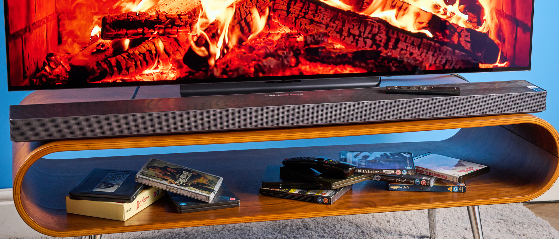 the samsung qs750f soundbar with two rear surround speakers and subwoofer photographed against a blue background and a TV
