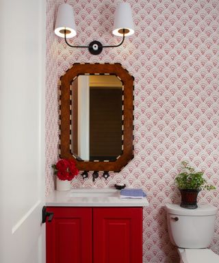 A pink bathroom with a light pink patterned wallpaper on the walls, a deep pink bathroom vanity topped with a white countertop, a wooden vanity mirror above, and a double iron wall light with white linen shades sat at the top of the mirror