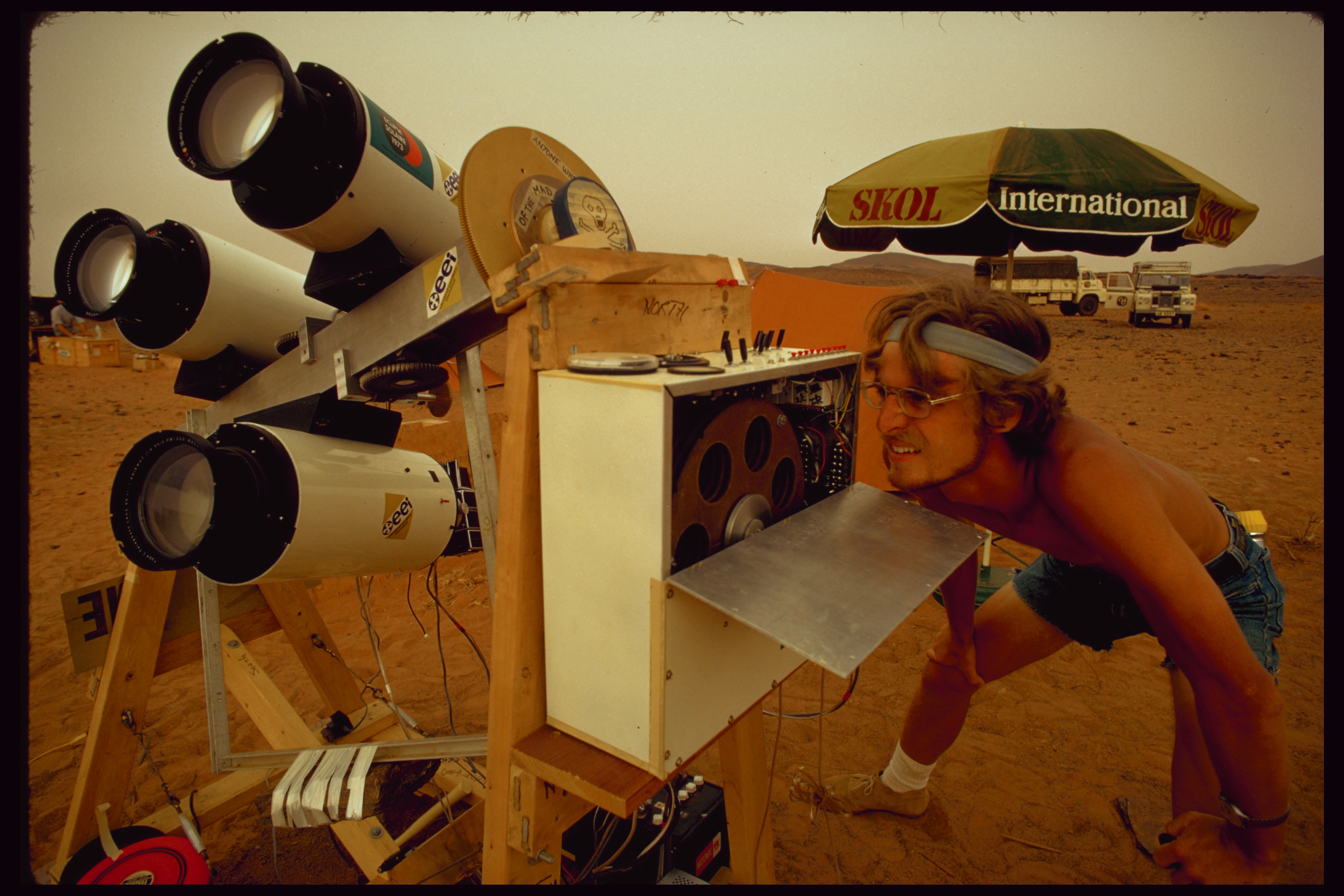 A man wearing denim shorts and no shirt inspects his telescope and viewing equipment.