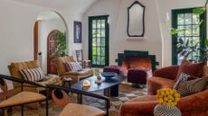 A white living room with forest green windows, a green fireplace, maroon seating stools, a curved copper sofa, a set of wooden chair, and two mustard sofa seaters by an oval coffee table and a checkerboard rug