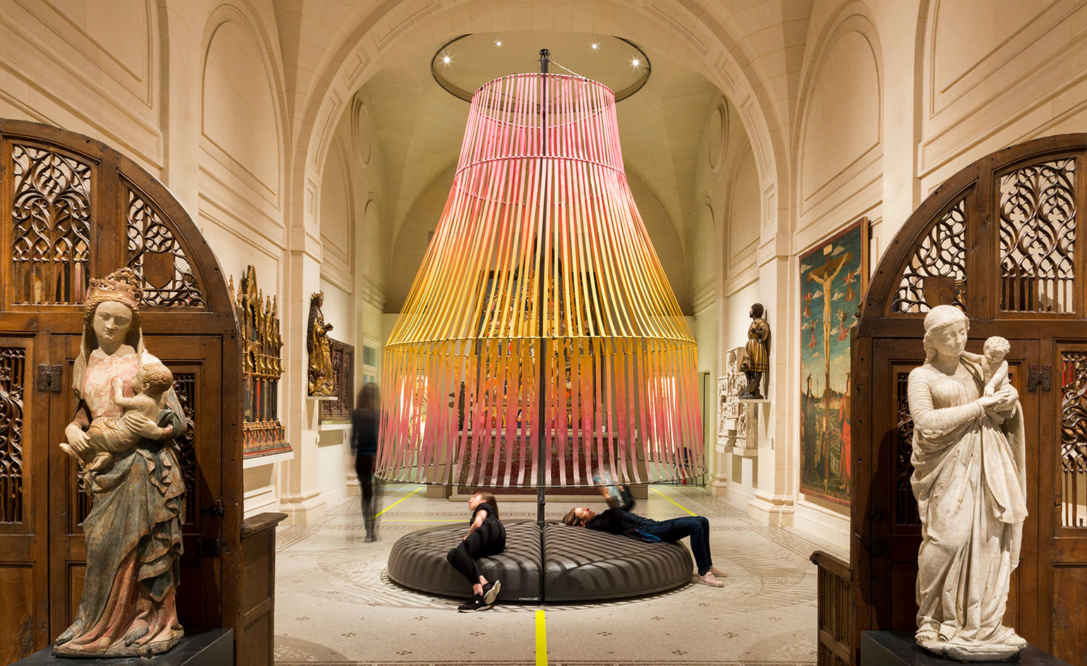 A room in a museum, with high ceilings, paintings, and sculptures placed throughout the room. A huge lamp-like art piece is set in the center, with seating below it.