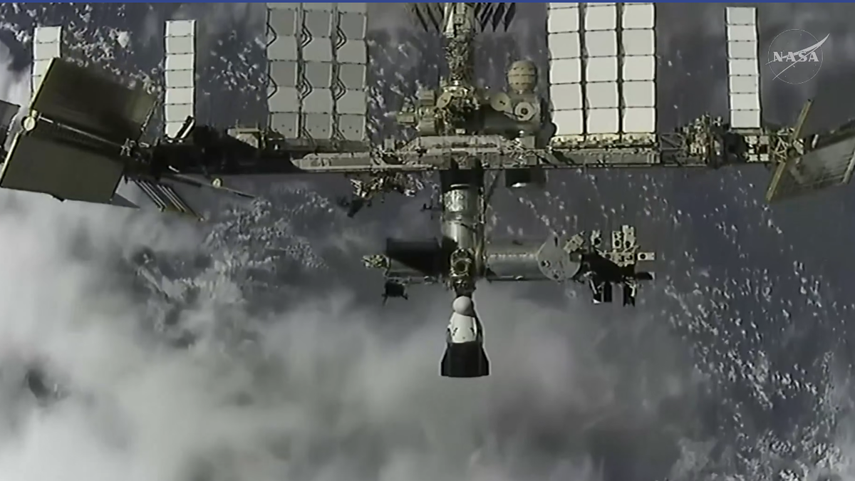 Black and white image looking down on the ISS with a dragon docked near the bottom