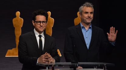 Two gentlemen is black suits standing at podium in front of images of 'Oscars' announcing nominations