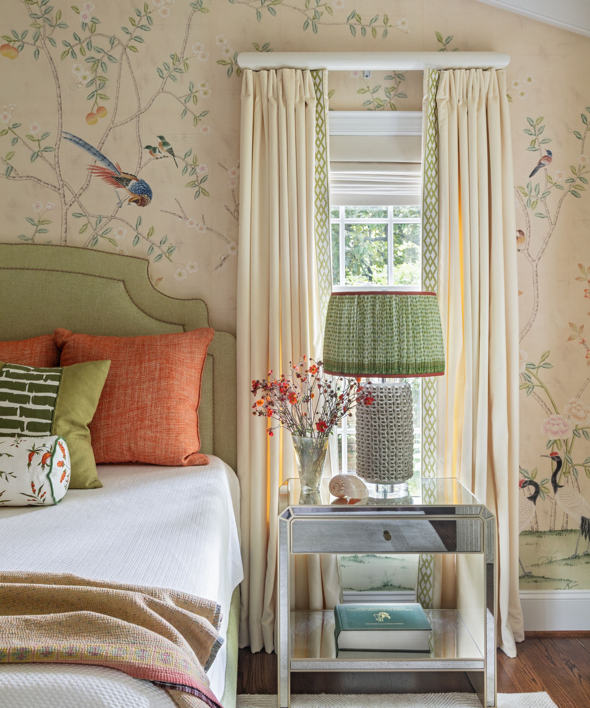 Bedroom with a bird motif wall mural with a pale peach background. Cream-colored drapes, a mirrored glass nightstand and a green lamp, and a green headboard with white bedding and orange cushions.