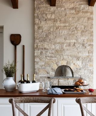 A kitchen with a light gray brick wall with a silver bread oven, a dark brown countertop with a tub of champagne bottles, glasses, and plates and cake stands with pastries, plus two dark wooden chairs in front
