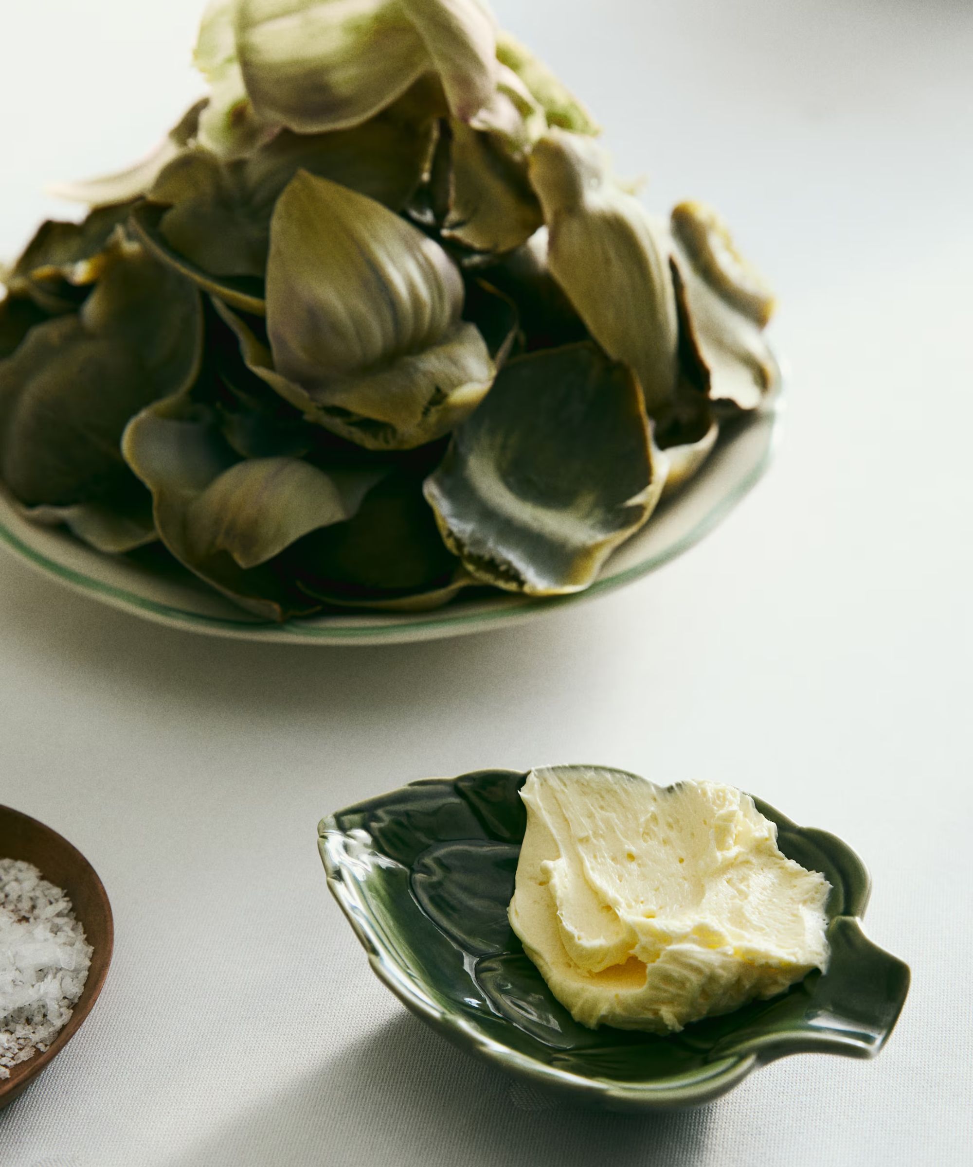 Artichoke shaped small dish next to green and white serving bowl filled with artichoke leaves