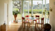 A bright dining room with floor to ceiling glass windows and a door leading to the backyard. In the dining room is a glass table with four wood chairs, pendant lighting above, a potted tree and a plant in a tall vase on a short plinth, a checkerboard rug and a wood table sculpted into an 'S'