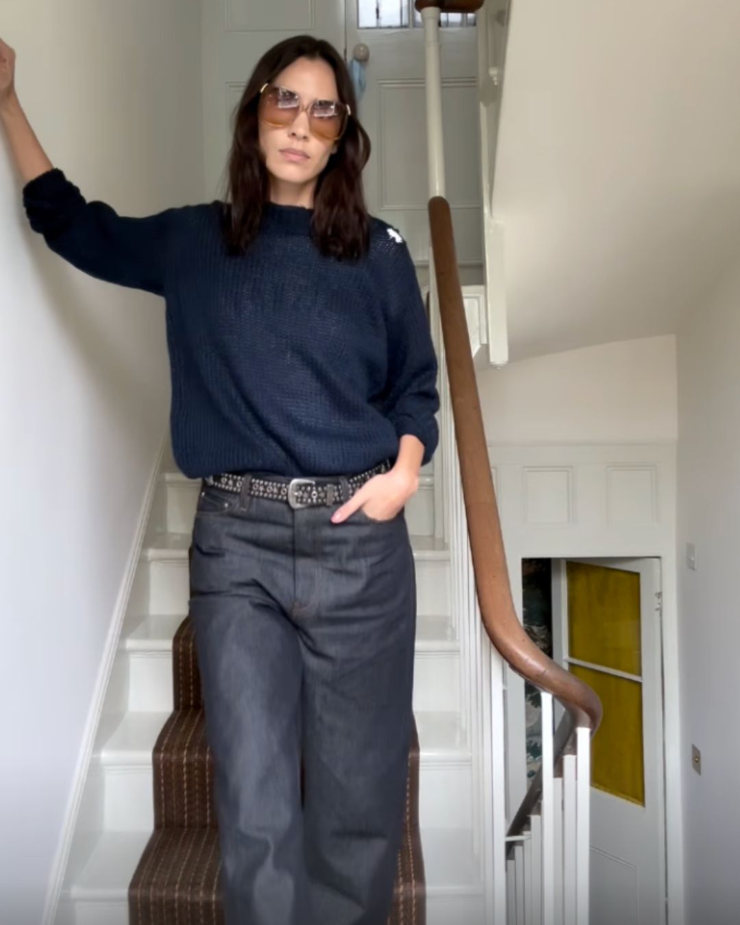 Alexa Chung stands on a staircase wearing a dark navy jumper tucked into indigo jeans. She wears a belt with her jeans and stands with her hand in her pocket.