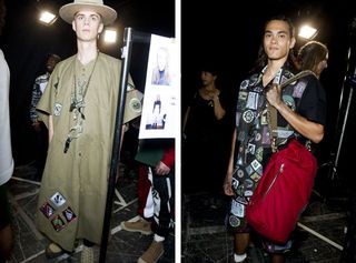 Guys wearing Man S/S 2015 collection. The guy on the left is wearing a green tent-like cape with totemic patches at the bottom and a cream hat. The guy on the right is wearing a short sleeve shirt filled with totemic images and a matching short with a red bag over his shoulder.