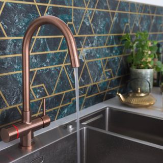Franke’s Minerva 4-in-1 Electronic tap in a copper colour with a gold teapot, potted plant and blue and gold wall tiles
