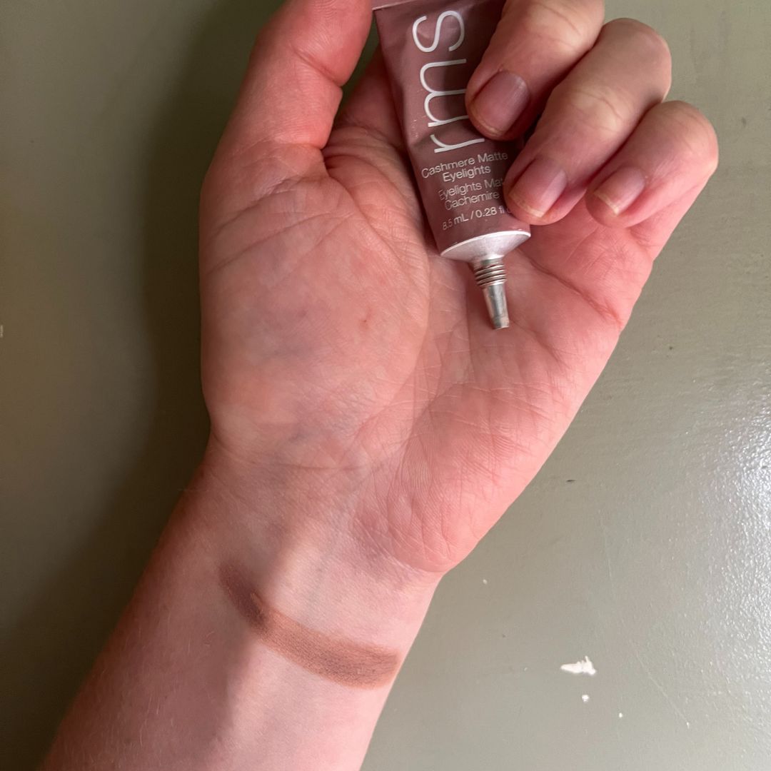 a hand on a green background holding RMS eyelights cream eyeshadow with a swatch of the eyeshadow on the wrist 
