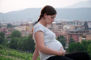 A pregnant woman sits on a hillside near a city.