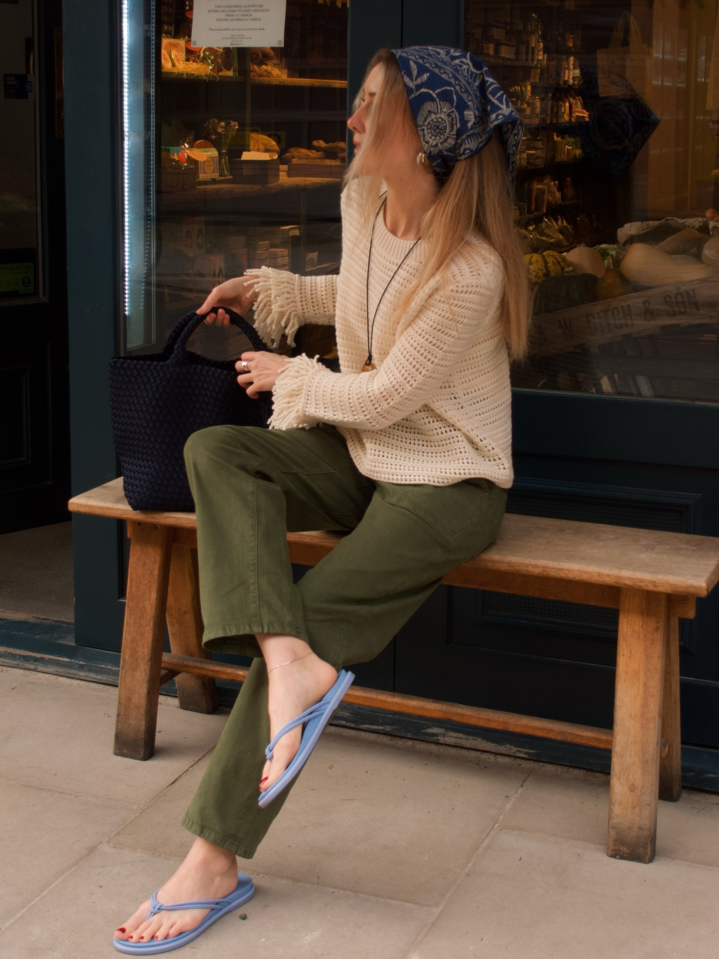 British style influencer poses on a bench outside a store in London wearing a blue printed bandana head scarf, an open-knit sweater, black tote bag, green pants, and light blue flip-flop sandals