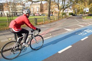 A commuter is shown in the image wearing a red jacket and helmet riding a bike on a blue bike lane travelling from left to right. In the back ground is a park and houses in the distance.
