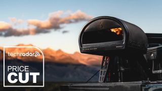 Gozney Roccbox pizza oven in black, sitting on the tailgate of a pickup truck, with mountains in the background. "Price cut" text in white is in the bottom left corner.