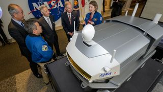 three men in suits and two astronauts wearing blue flight suits stand beside a model of a moon rover