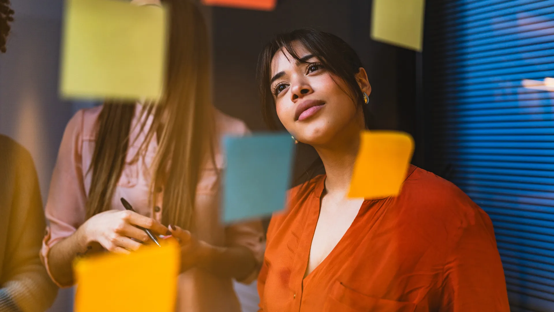 Businesswoman in an orange shirt thoughtfully reading notes on a glass wall during a brainstorming session, highlighting collaboration and strategic thinking in a modern office