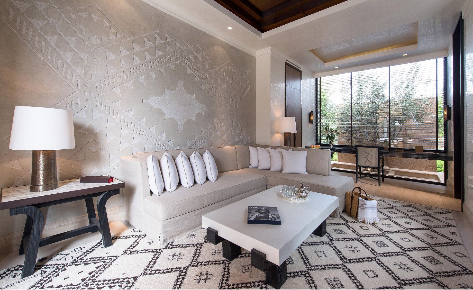 A hotel lounge area with a corner sofa, a long desk, a square coffee table, a wooden console, patterned floor tiles and large glass windows.