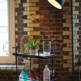 A bar trolley with a tall floor lamp against an exposed brick wall