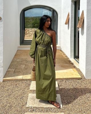 British influencer Monikh Dale poses outside wearing a one-shoulder olive green dress from Faithfull the Brand, a small basket bag, and strappy flat sandals.