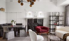 A reading room with off-white walls, grey rug, grey marble accents, white curved sofa and plum armchair, plus dining area with round black wood pedestal dining table, corner banquette upholstered in black fabric, and grey and white patterned chairs