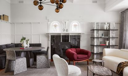 A reading room with off-white walls, grey rug, grey marble accents, white curved sofa and plum armchair, plus dining area with round black wood pedestal dining table, corner banquette upholstered in black fabric, and grey and white patterned chairs