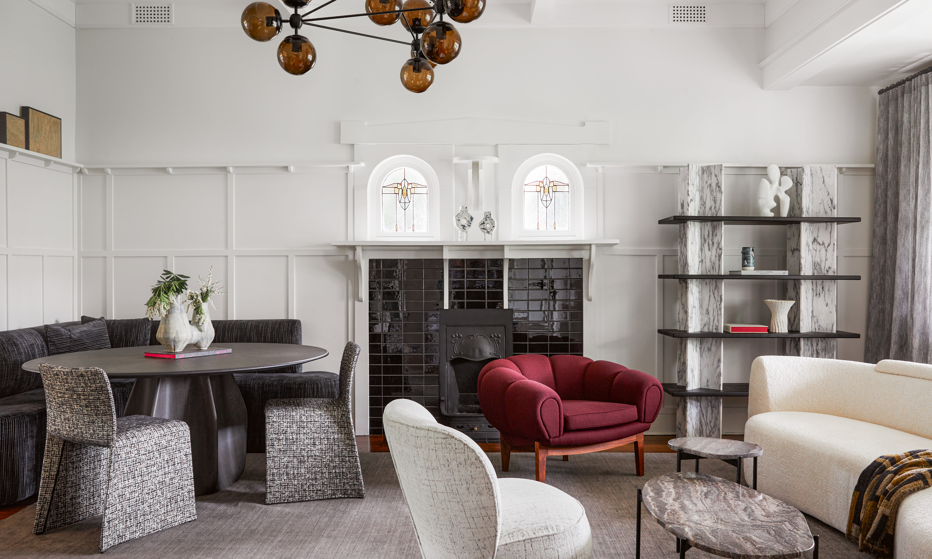 A reading room with off-white walls, grey rug, grey marble accents, white curved sofa and plum armchair, plus dining area with round black wood pedestal dining table, corner banquette upholstered in black fabric, and grey and white patterned chairs