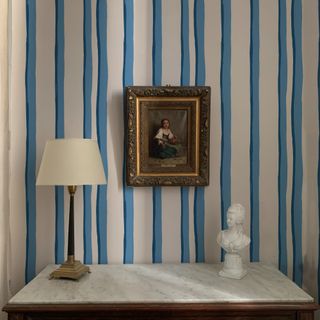 A console table with a table lamp and a bust with the wall behind covered in a striped wallpaper and bearing a framed artwork