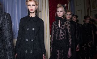Six female models wearing looks from Simone Rocha's collection. One model is wearing a black textured coat with buttons. Another model is wearing a black sheer dress with embellishments and has a bow at the neck. And the other models are wearing dark coloured pieces