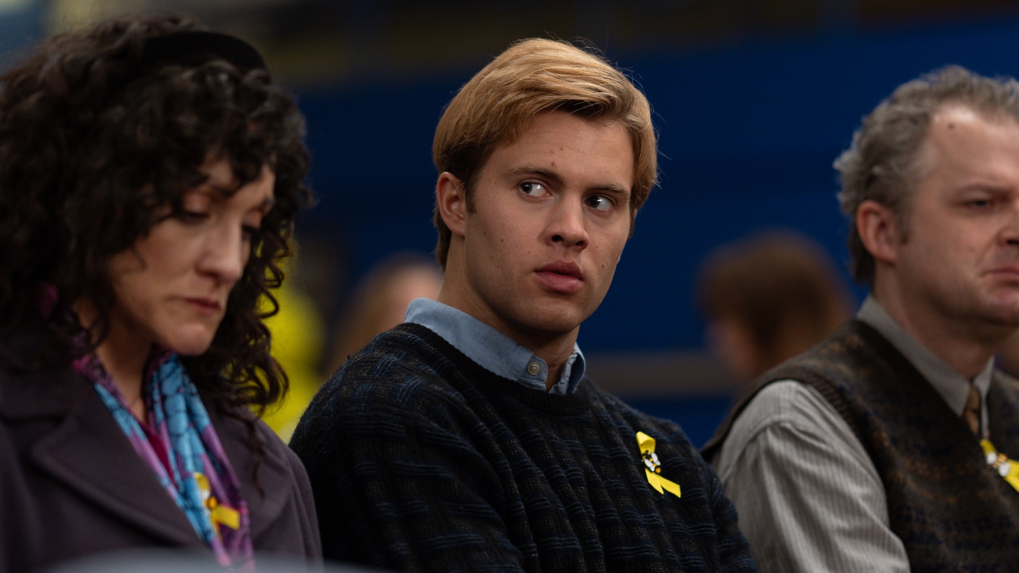 Teen Jeff swearing sweater with yellow ribbon pinned to it in between two adults in Yellowjackets Season 4