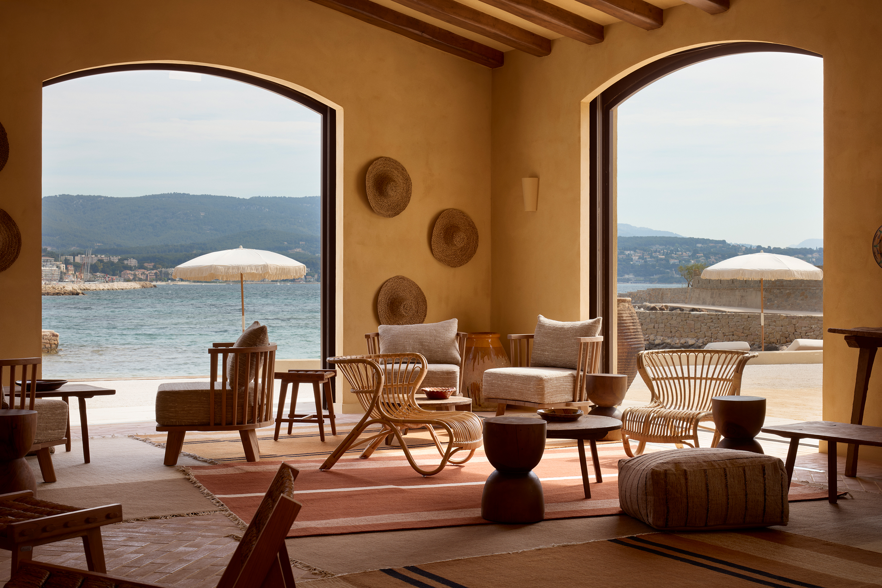 The ochre-tinted interiors of a sea-view bar, decorated with rustic wooden furniture, textural fabrics, and earthy tones throughout, with arched floor-to-ceiling windows.