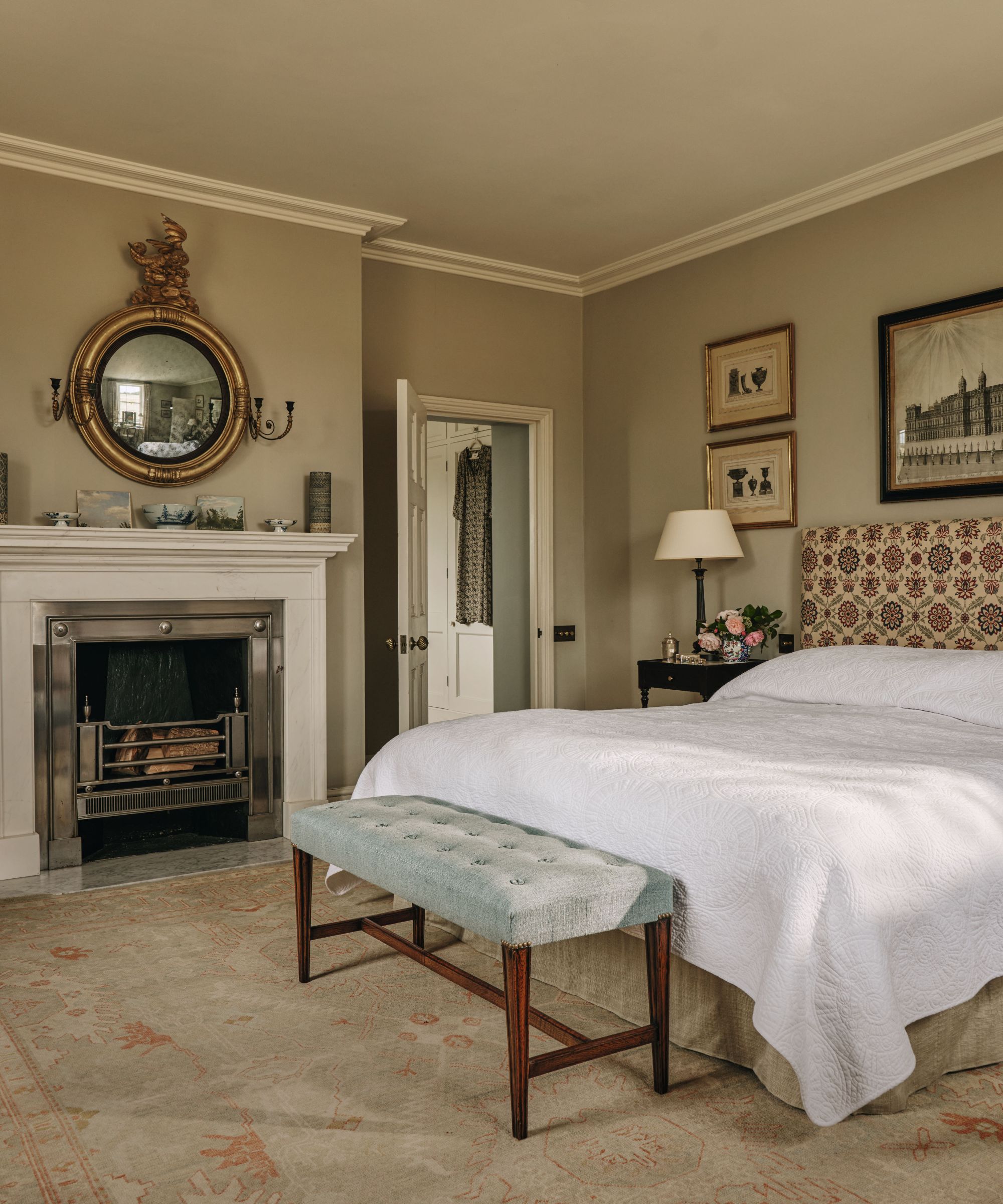 Elegant bedroom with a patterned upholstered headboard, white bedspread, a blue bench at the foot of the bed, and a classic fireplace
