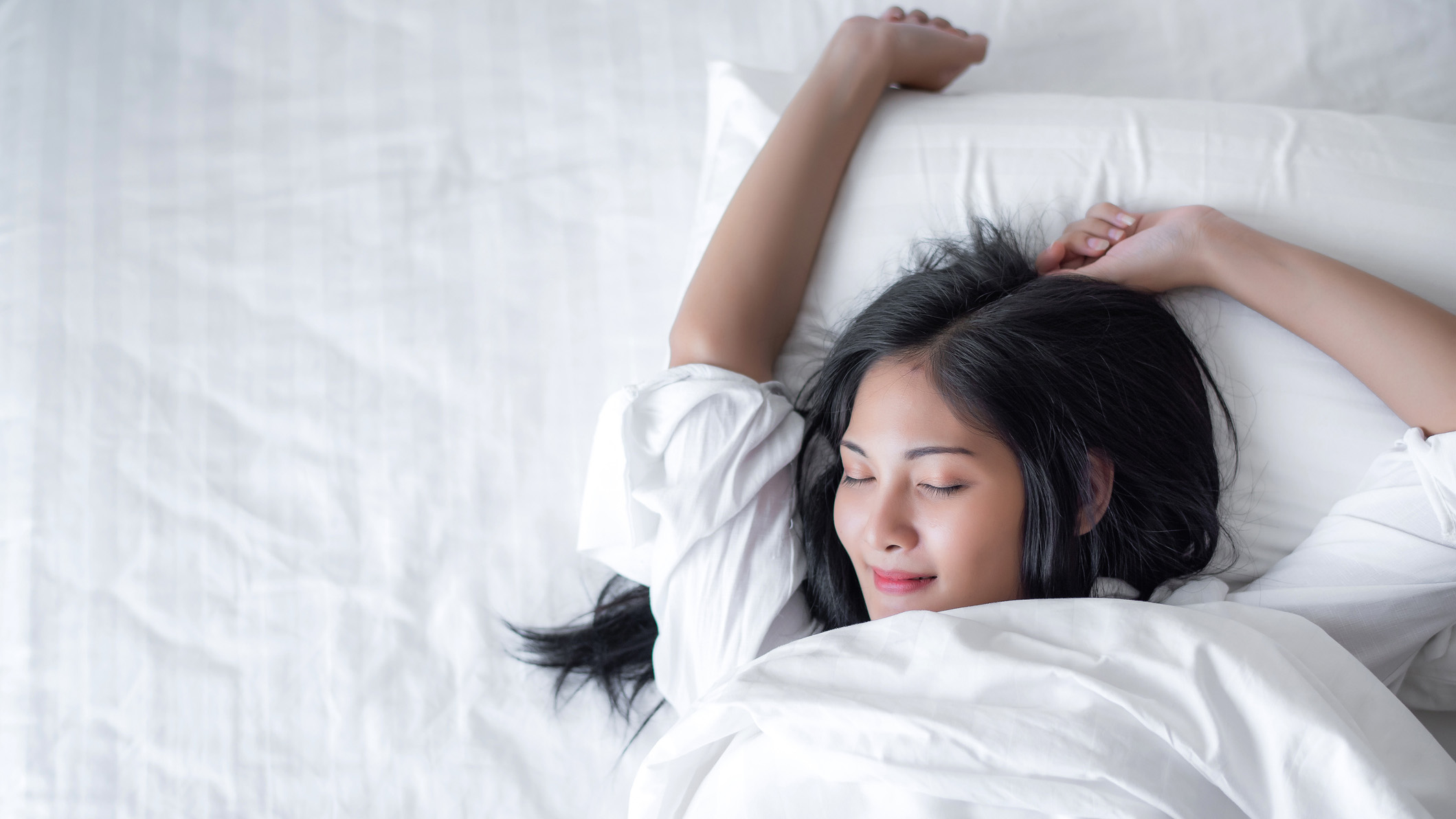 A woman sleeps happily with her arm raised on a comfortable white mattress