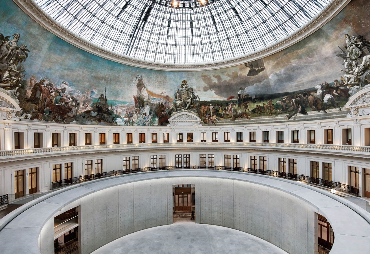 The Bourse de Commerce in Paris. Large, circular hall, with renaissance art on the walls and a glass dome.