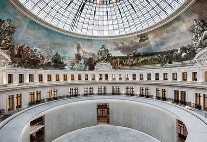 The Bourse de Commerce in Paris. Large, circular hall, with renaissance art on the walls and a glass dome.