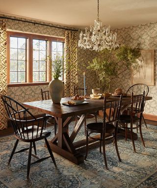 A dark wood dining set on a blue and beige rug; on the walls, green floral wallpaper; hanging from the ceiling, a chandelier.