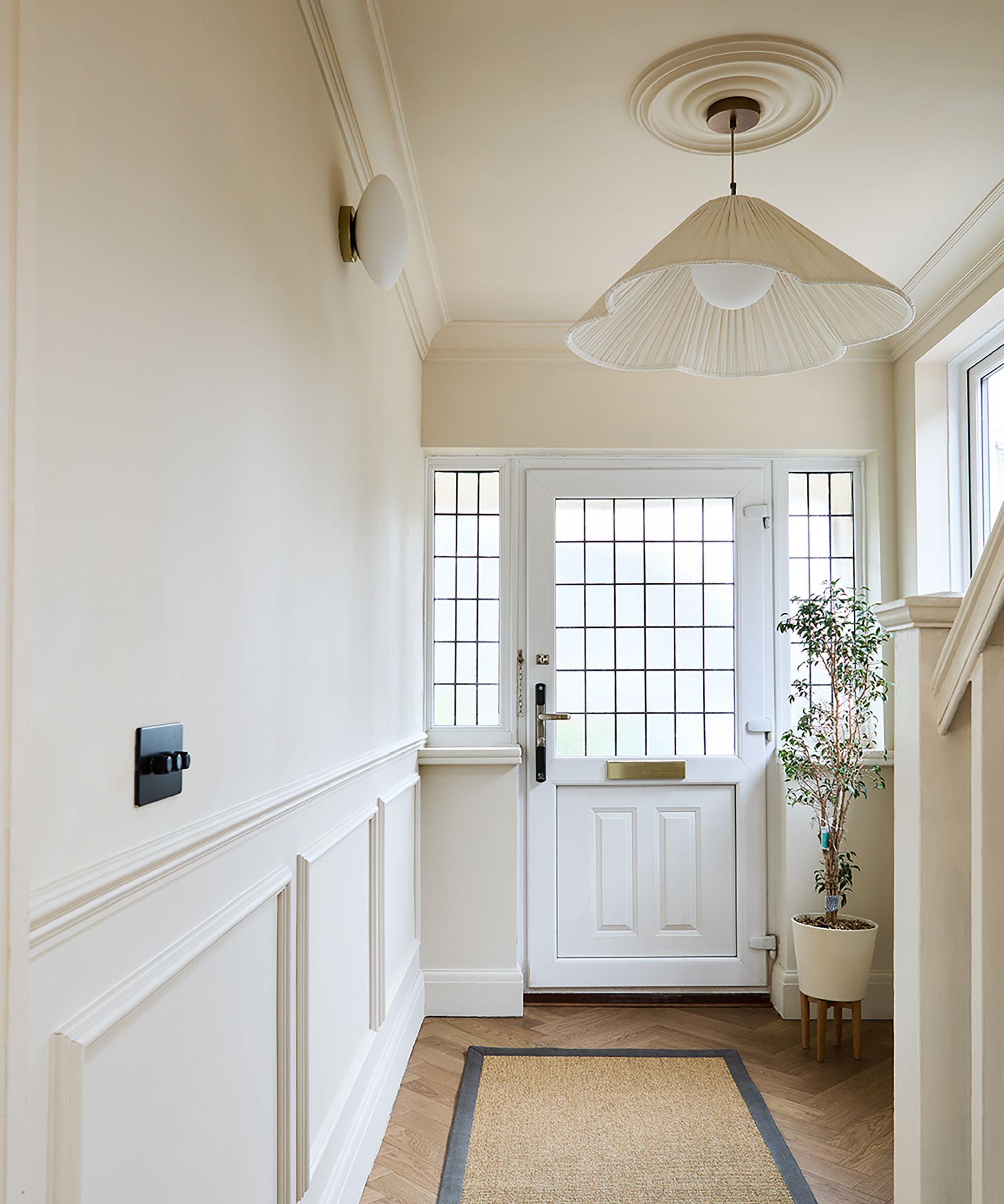Crisp hallway with white panelled walls, geometric light fitting and grid-glazed front door, finished with warm herringbone flooring.