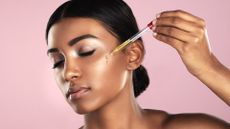 Studio shot of a beautiful young woman applying essential oil to her face with a dropper posing against a pink background