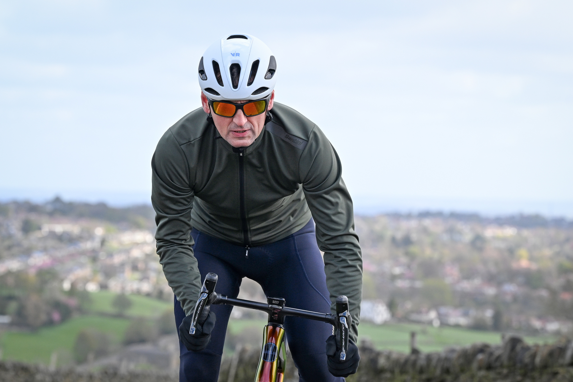Man wearing a dark green jacket, black gloves and white helmet riding towards the camera out of the saddle