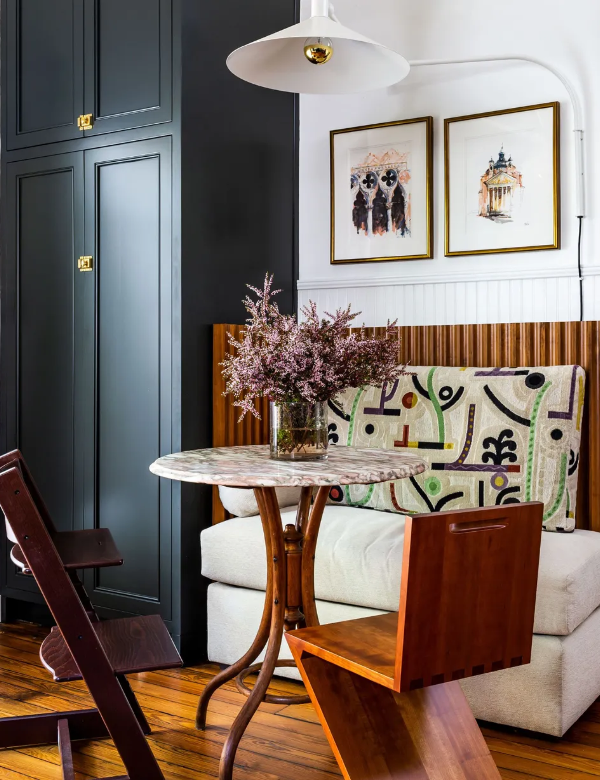 A breakfast nook featuring a marble-topped pedestal table, a modern Z-shaped wood chair, and a cream banquette with a vibrant, abstract-patterned cushion. A tall matte black cabinet and framed sketches on a white wall complete the corner