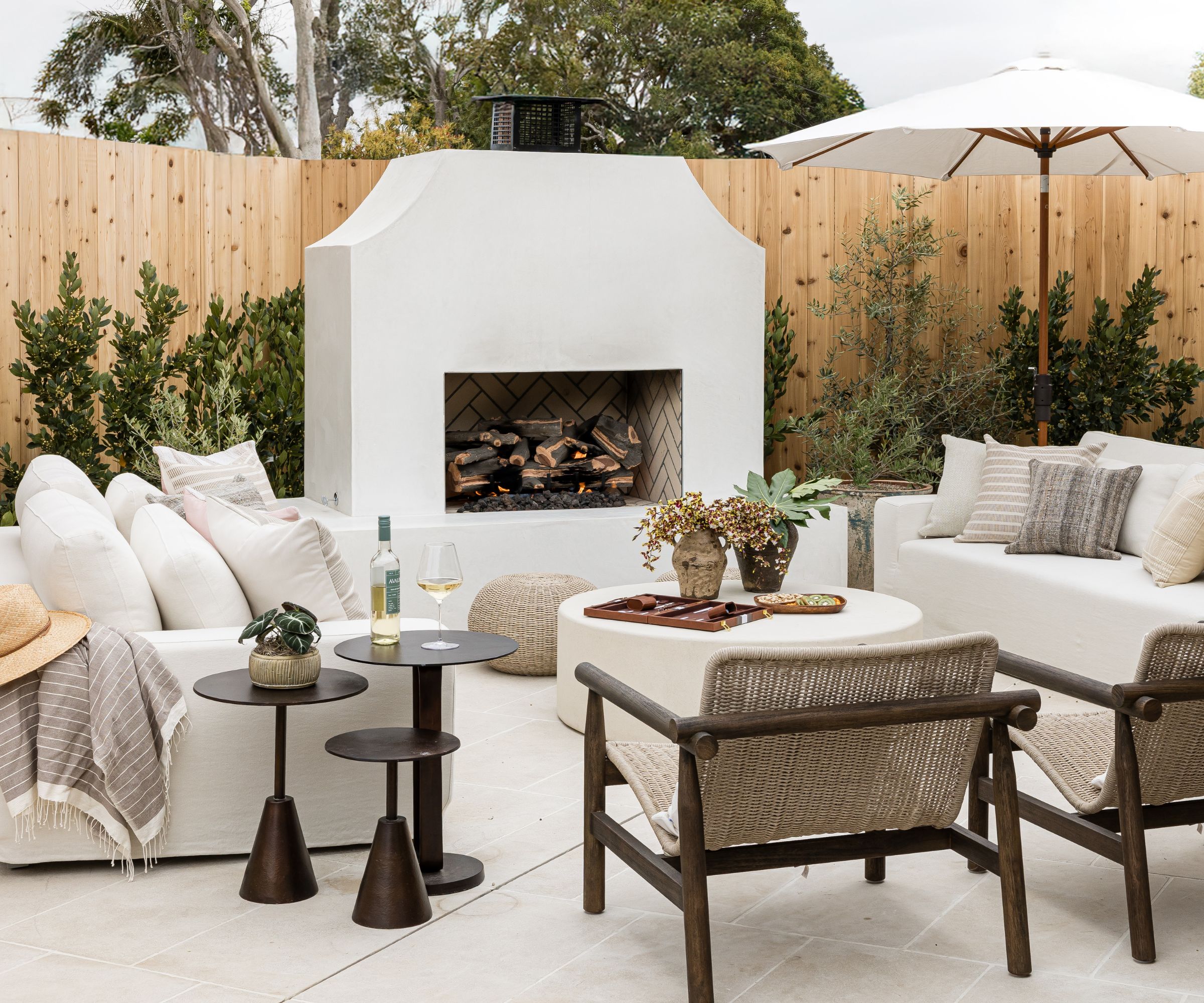 modern boho outdoor living room area designed around a white concrete fireplace, surrounded by plants with two sofas, armchairs and coffee tables