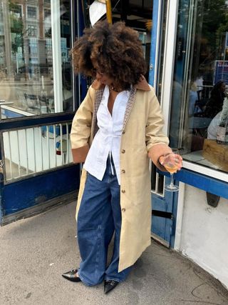 Amaka Hamelijnck with a voluminous afro wearing an oversize trench, white top, jeans, and heels.